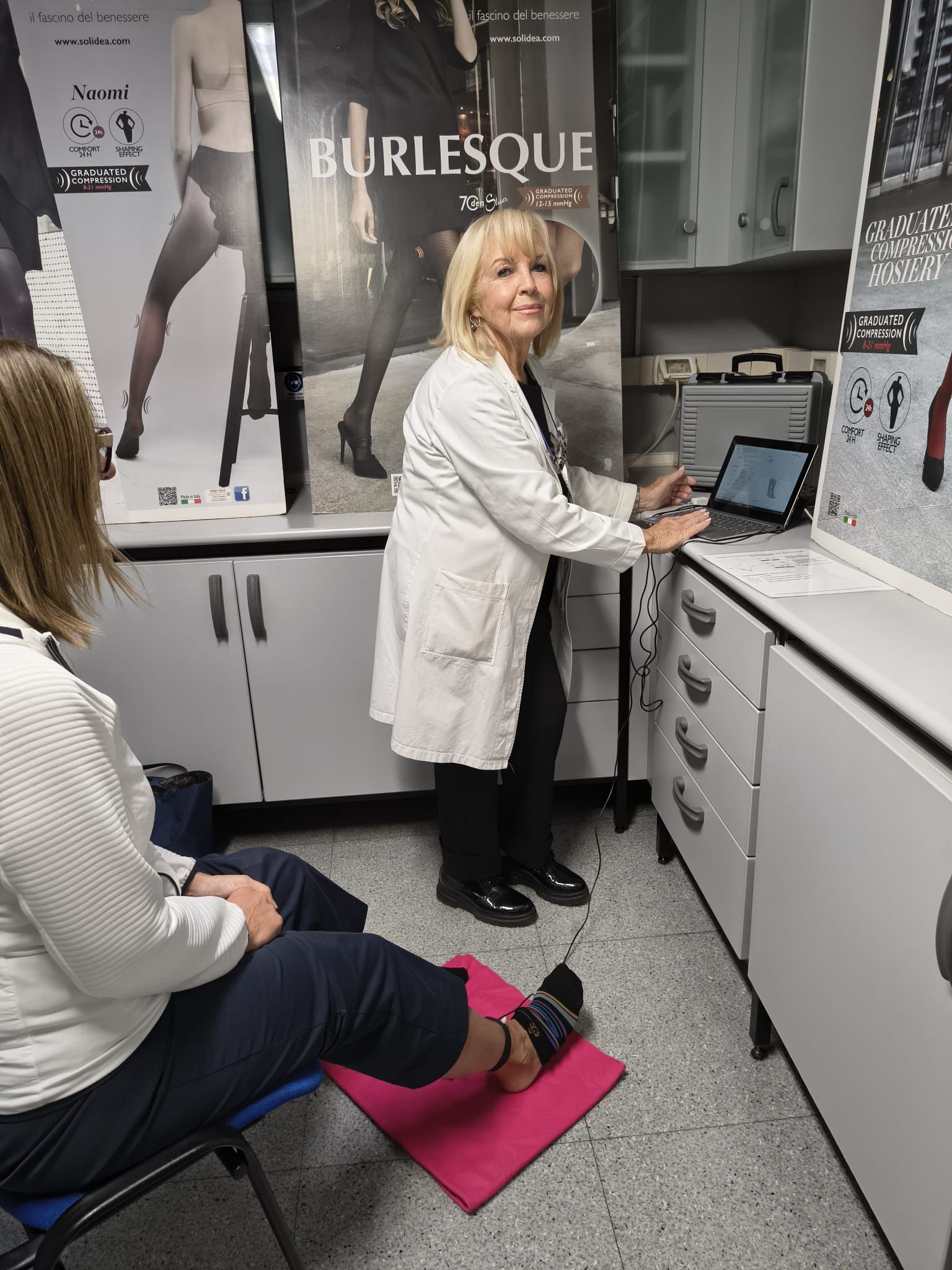 Una donna in camice da laboratorio aiuta un'altra donna a fare esercizi per le gambe, utilizzando un tablet.