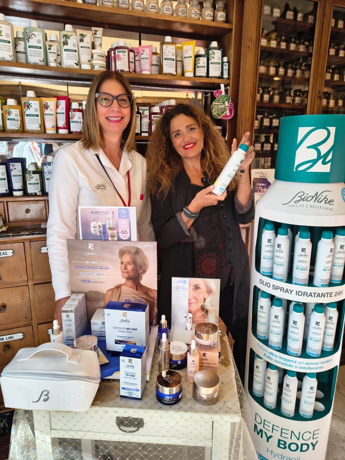 Due donne espongono prodotti per la cura della pelle in un negozio, una delle quali ne tiene in mano una bottiglia, con un espositore lì vicino.