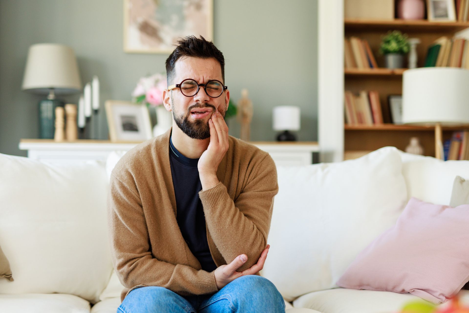 Man with a beard sits on a couch, holding his jaw in pain; indoor home setting.