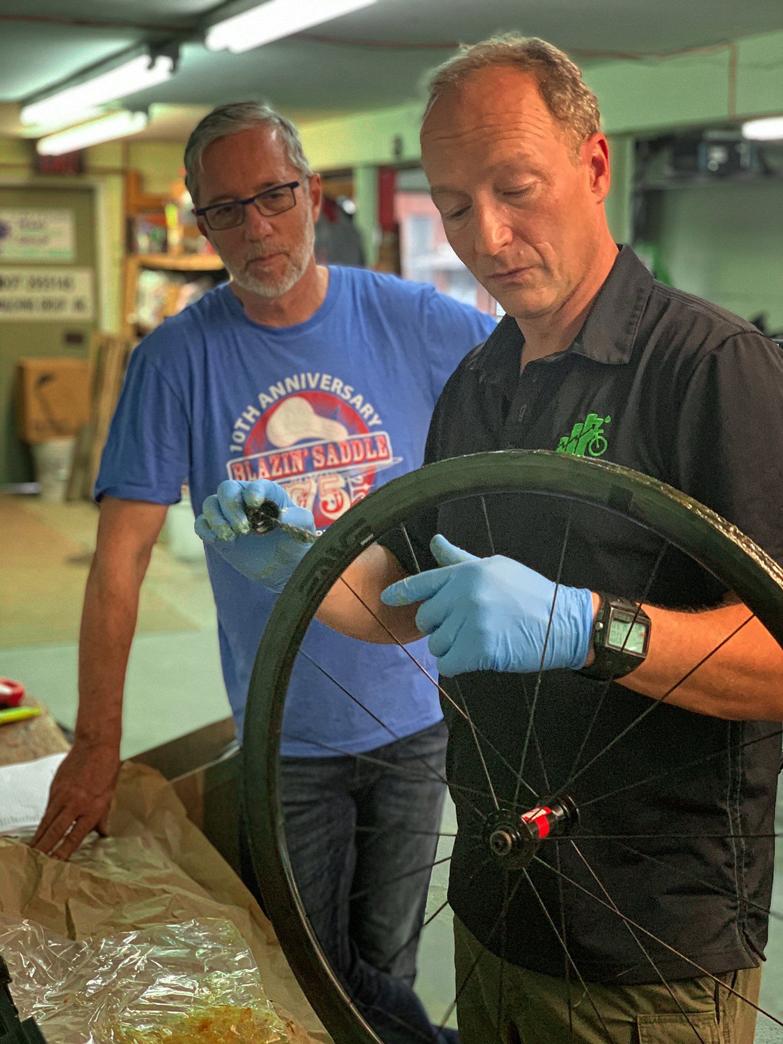 Two men are working on a bicycle wheel in a garage.