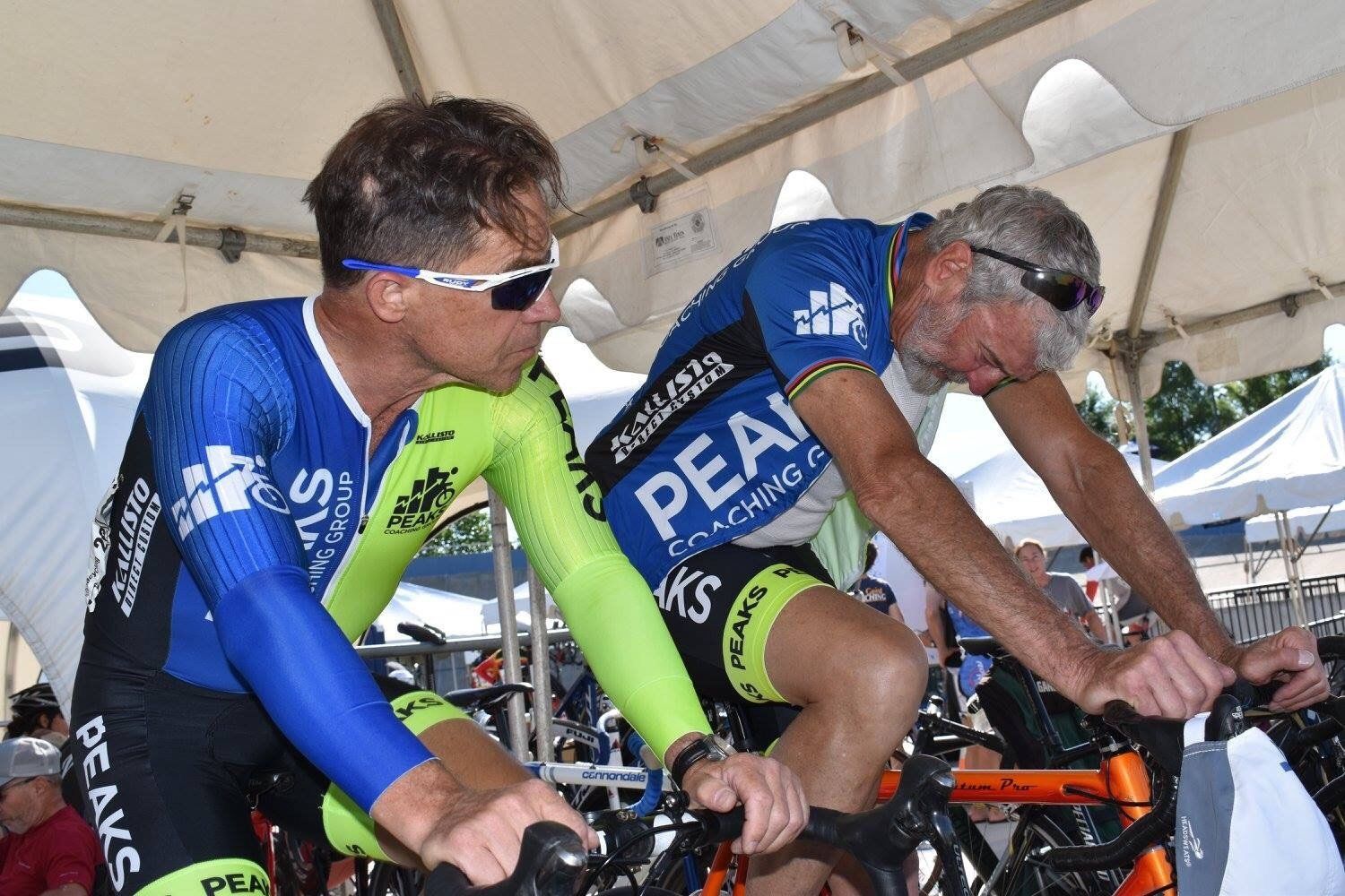 Two men are riding exercise bikes under a tent.
