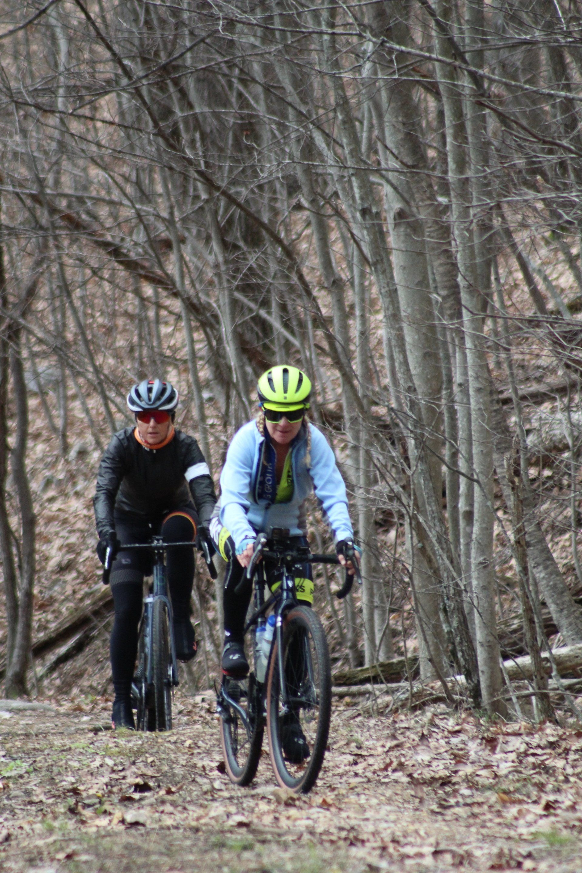 Two people are riding bicycles down a trail in the woods.