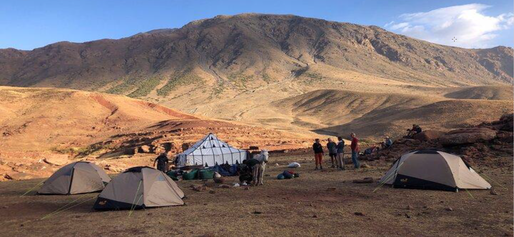 A group of people are standing around tents in the desert with mountains in the background.