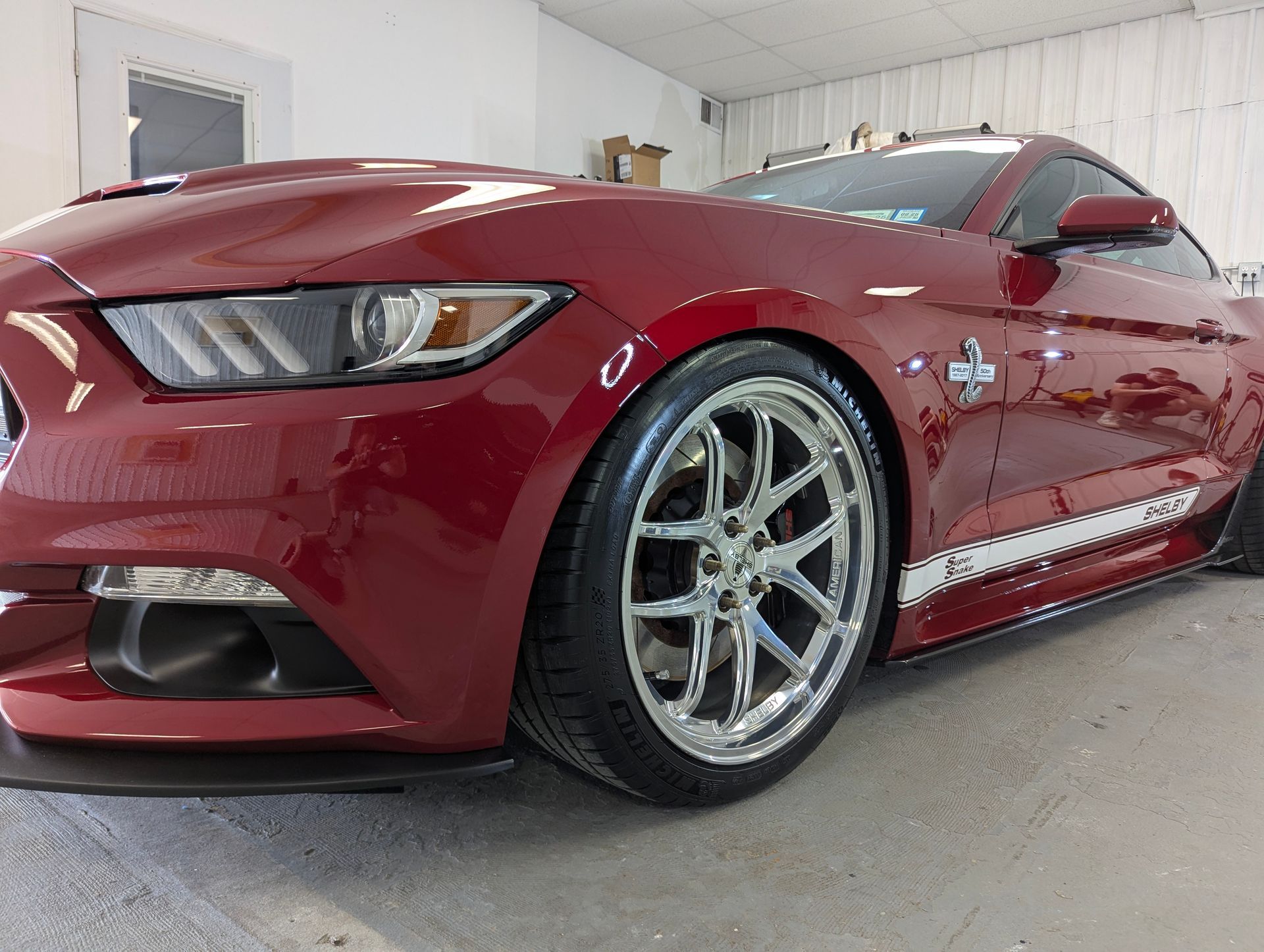 Red Shelby Mustang sports car, shiny chrome wheels, white racing stripes.