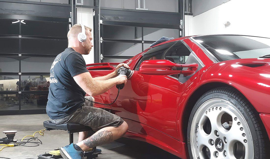 A man is sitting on a stool polishing a red sports car.