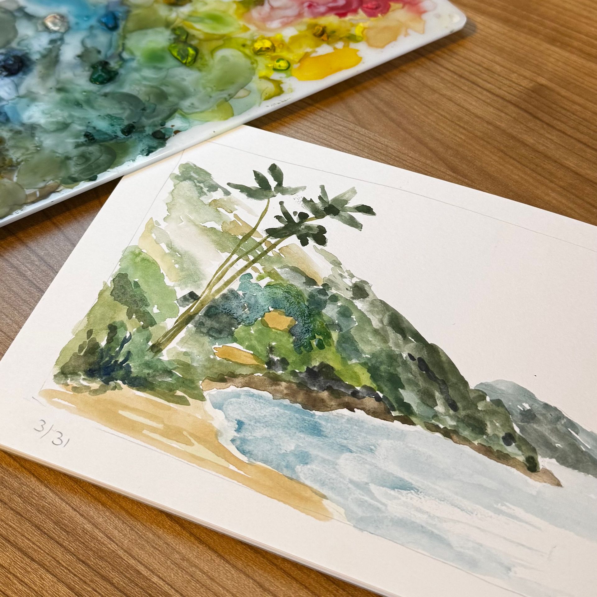 Aquarel van een strandtafereel met een berg, palmbomen en een palet met verf op een houten tafel.