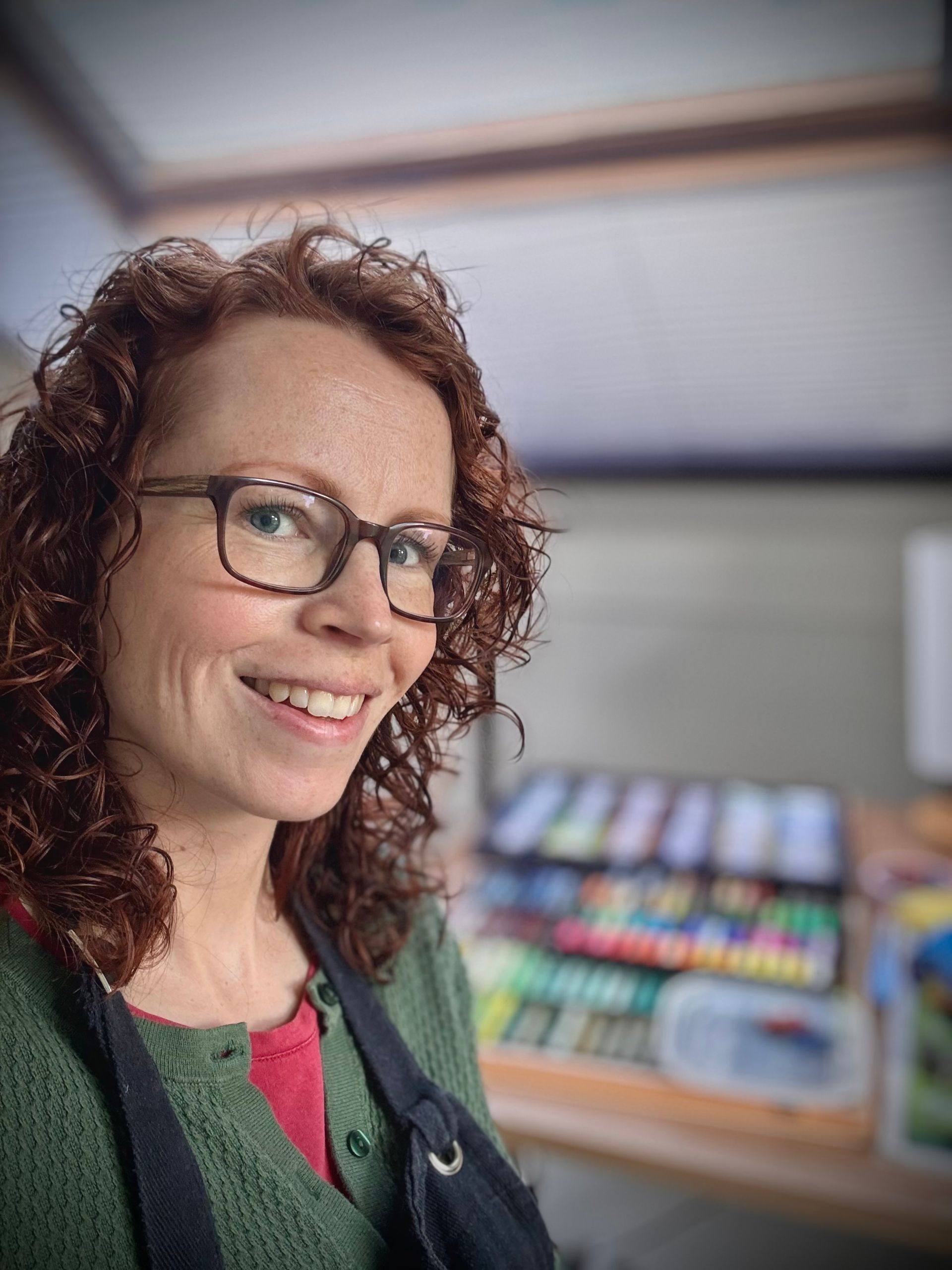 Een vrouw met krullend rood haar en een bril glimlacht en leunt naar de camera, pasteltinten op de achtergrond.