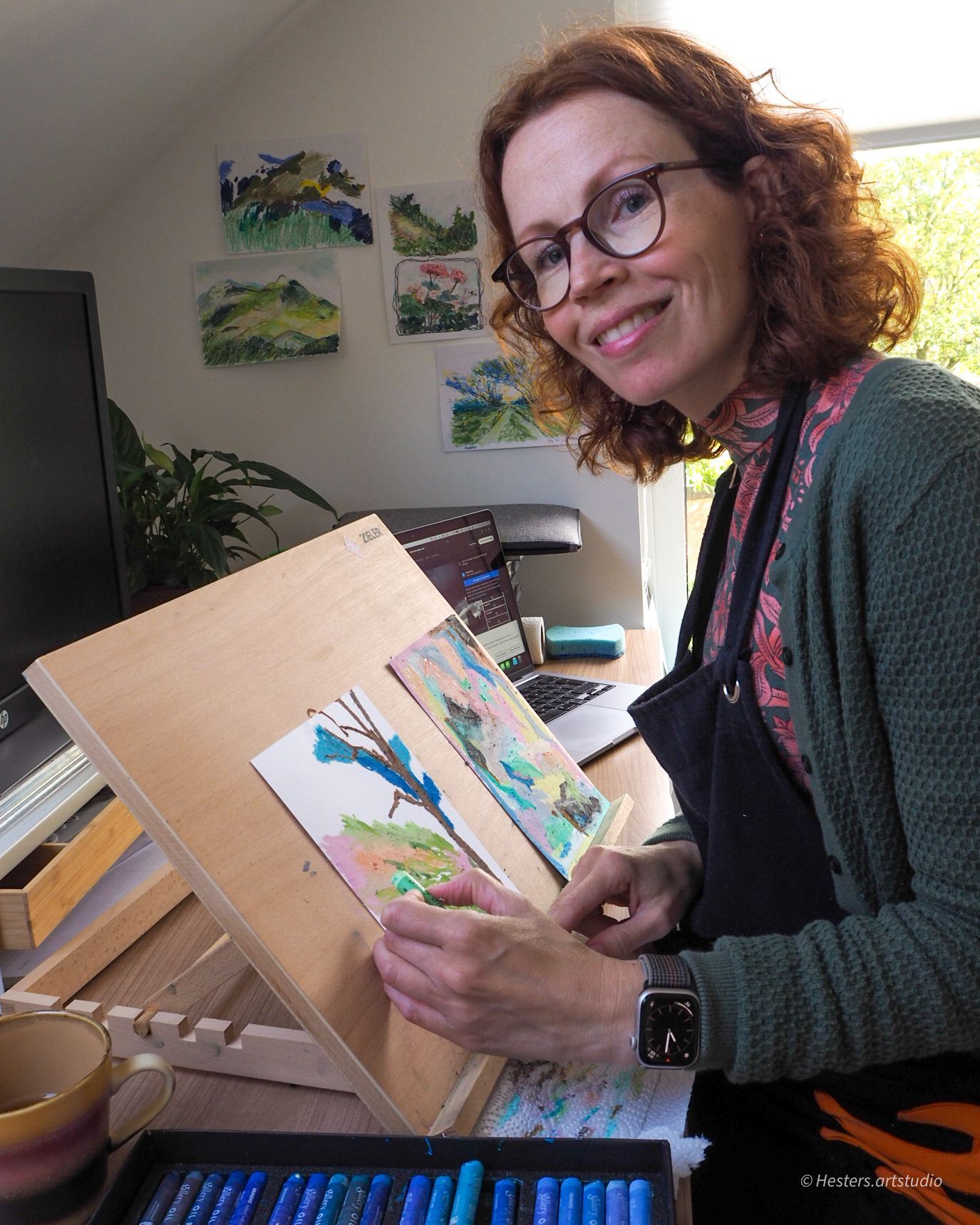 Vrouw aan een bureau met laptop, aquarelpalet en kleurstalen, aan het schilderen.