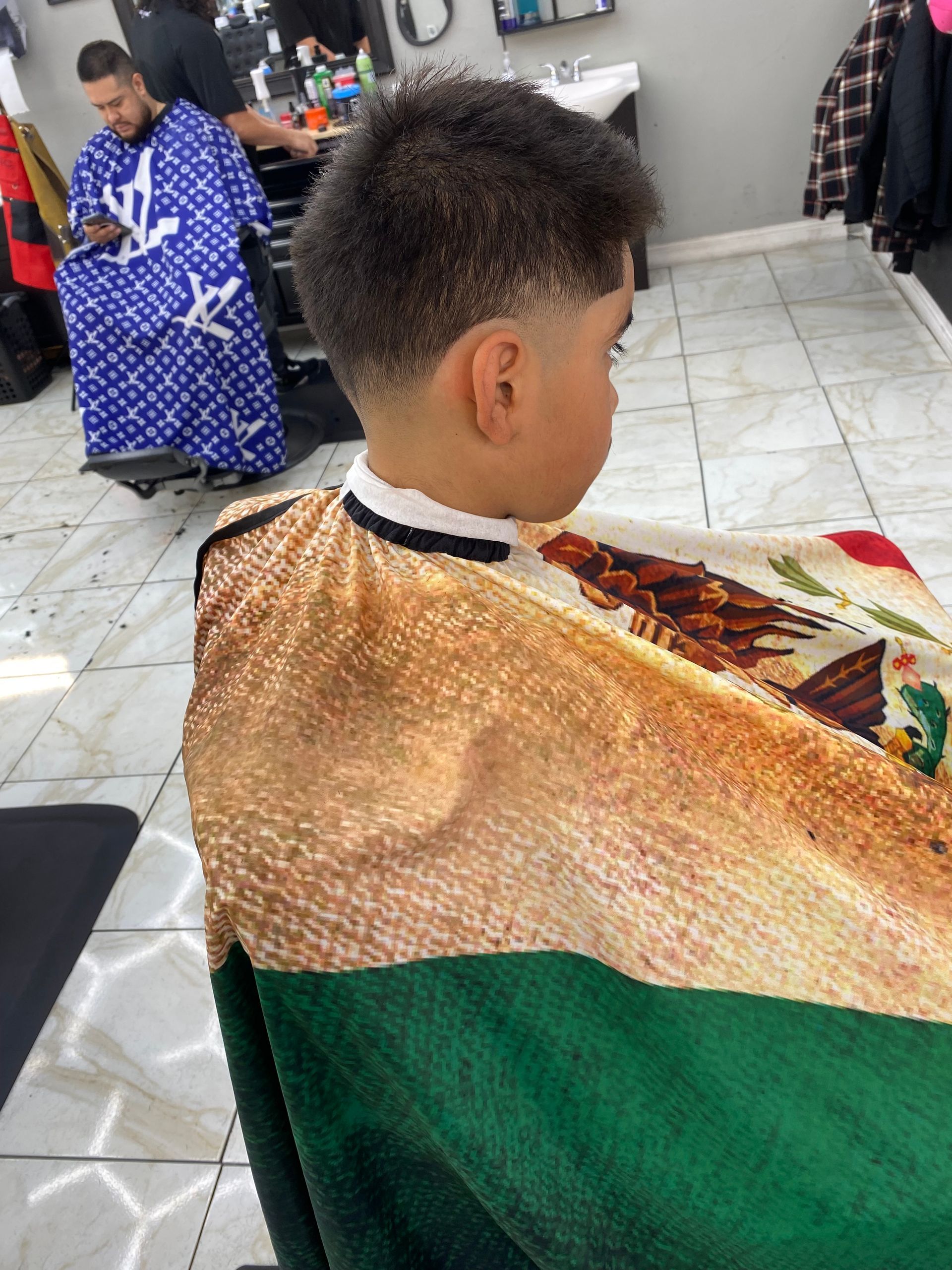 Boy in barber chair with a faded haircut, wearing a barber cape. Barber in background.