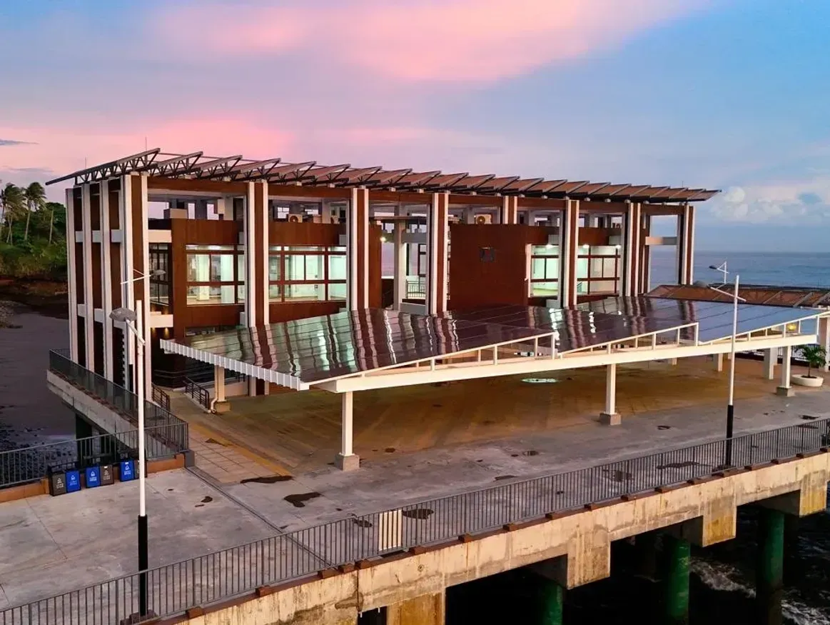 Una persona con cabello oscuro y rizado y una expresión cálida y sonriente sobre un fondo neutro de color blanco roto.