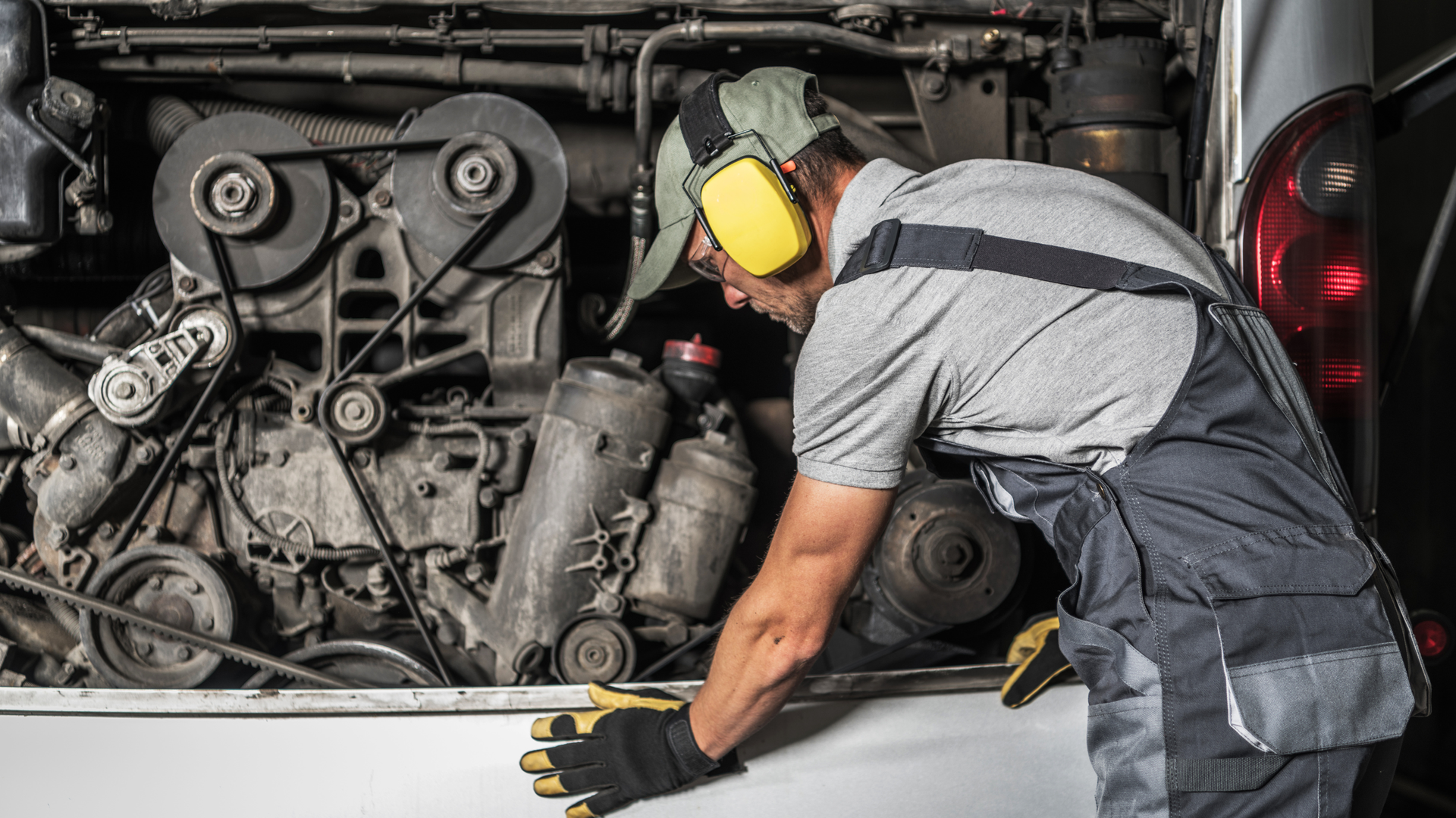 Mechanic in gray overalls and yellow ear protection inspecting a truck engine under the hood