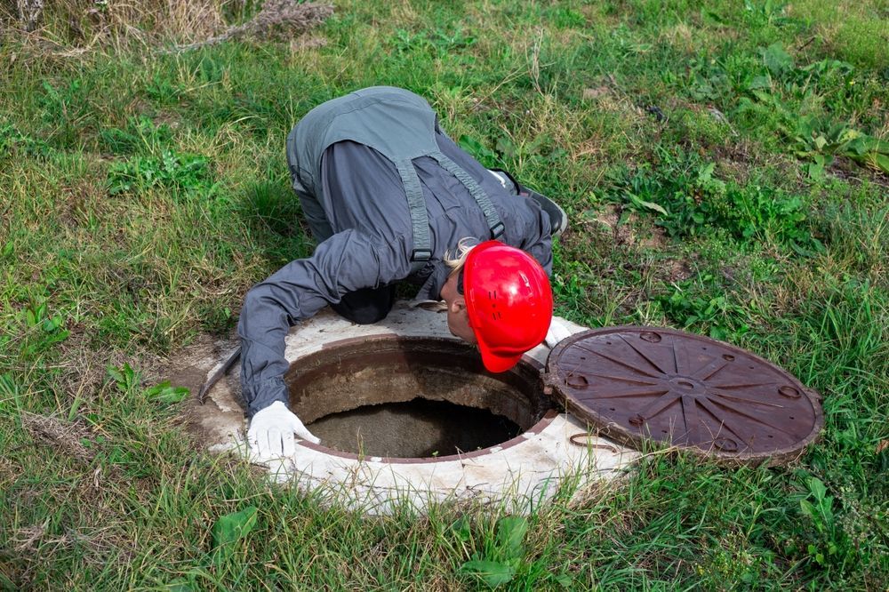 A man in a red hard hat is looking into a manhole cover.