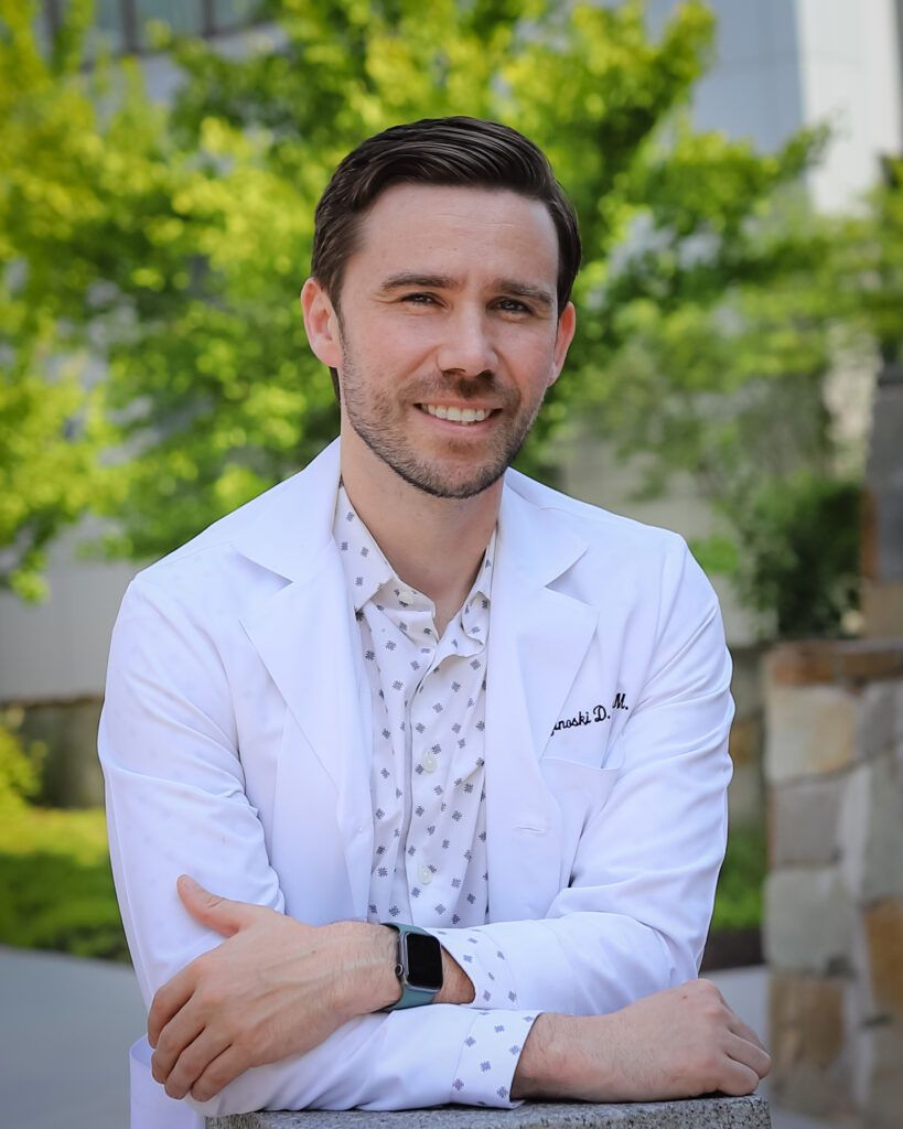 Man in white coat smiles, arms crossed, leaning on stone structure outdoors.