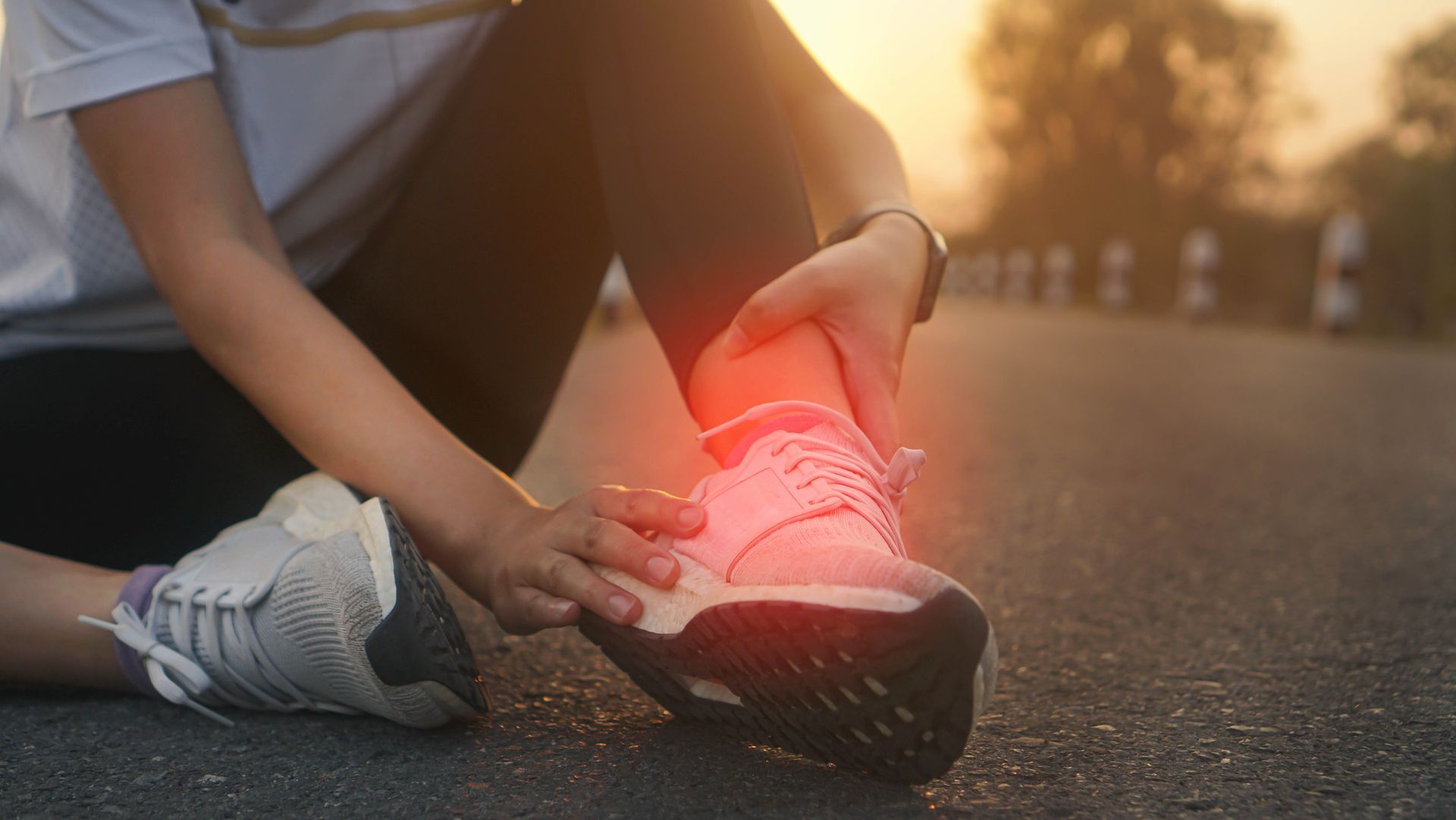 Person holding their ankle, indicating pain, while sitting on a road wearing athletic shoes; red glow highlights injury.