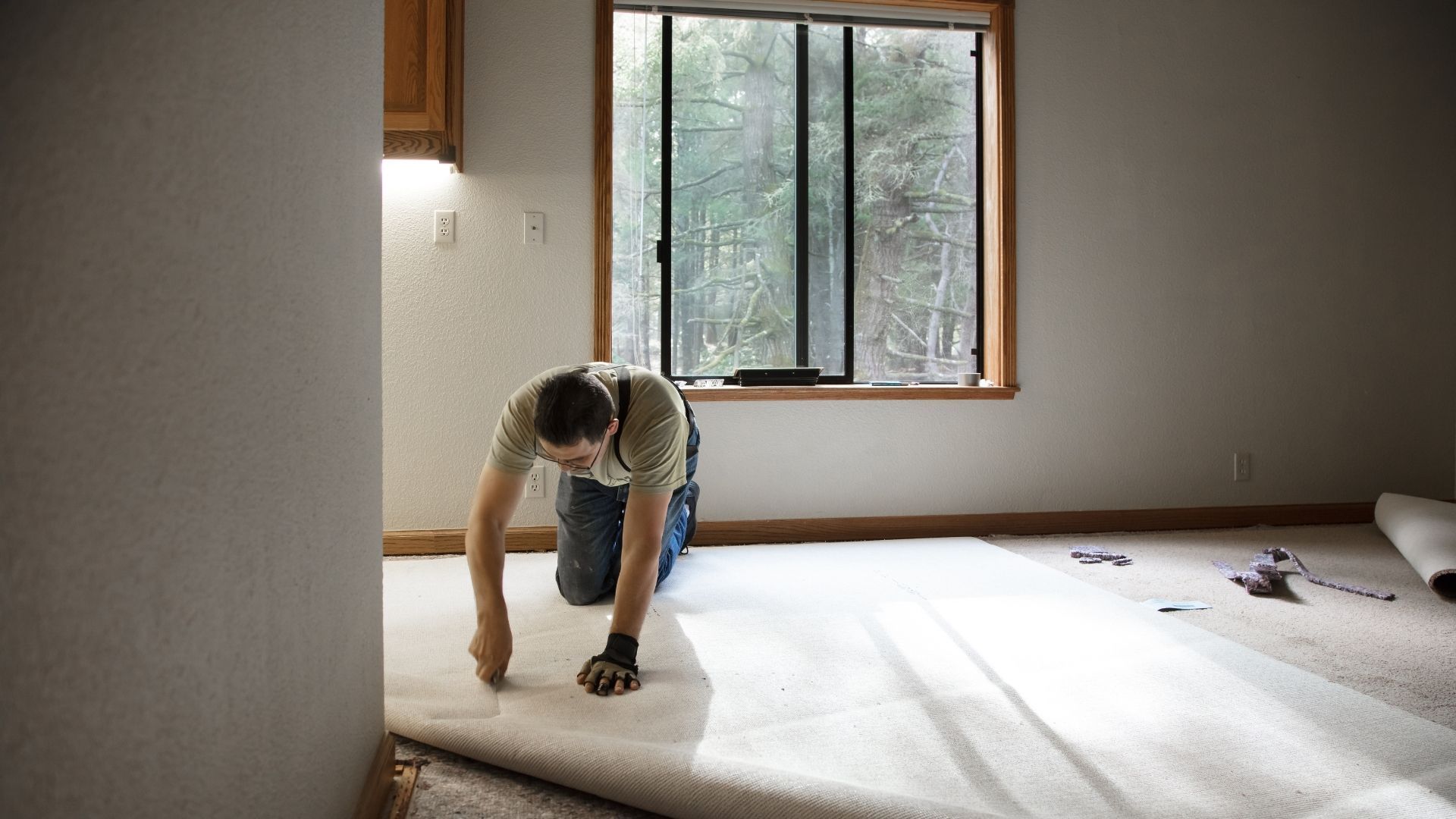 Man kneels, installing carpet in a room with a window and wooden cabinets.