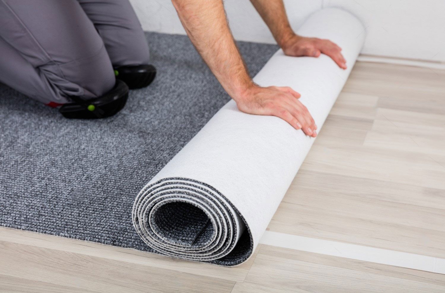 Person rolling out a new gray carpet on a light wooden floor.