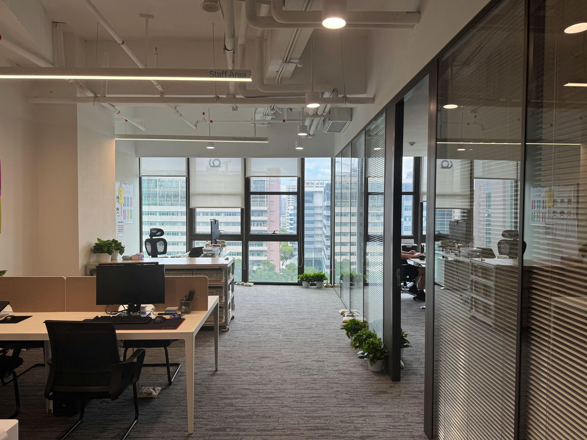 Office interior with desks, computers, glass walls, and a view of city buildings.