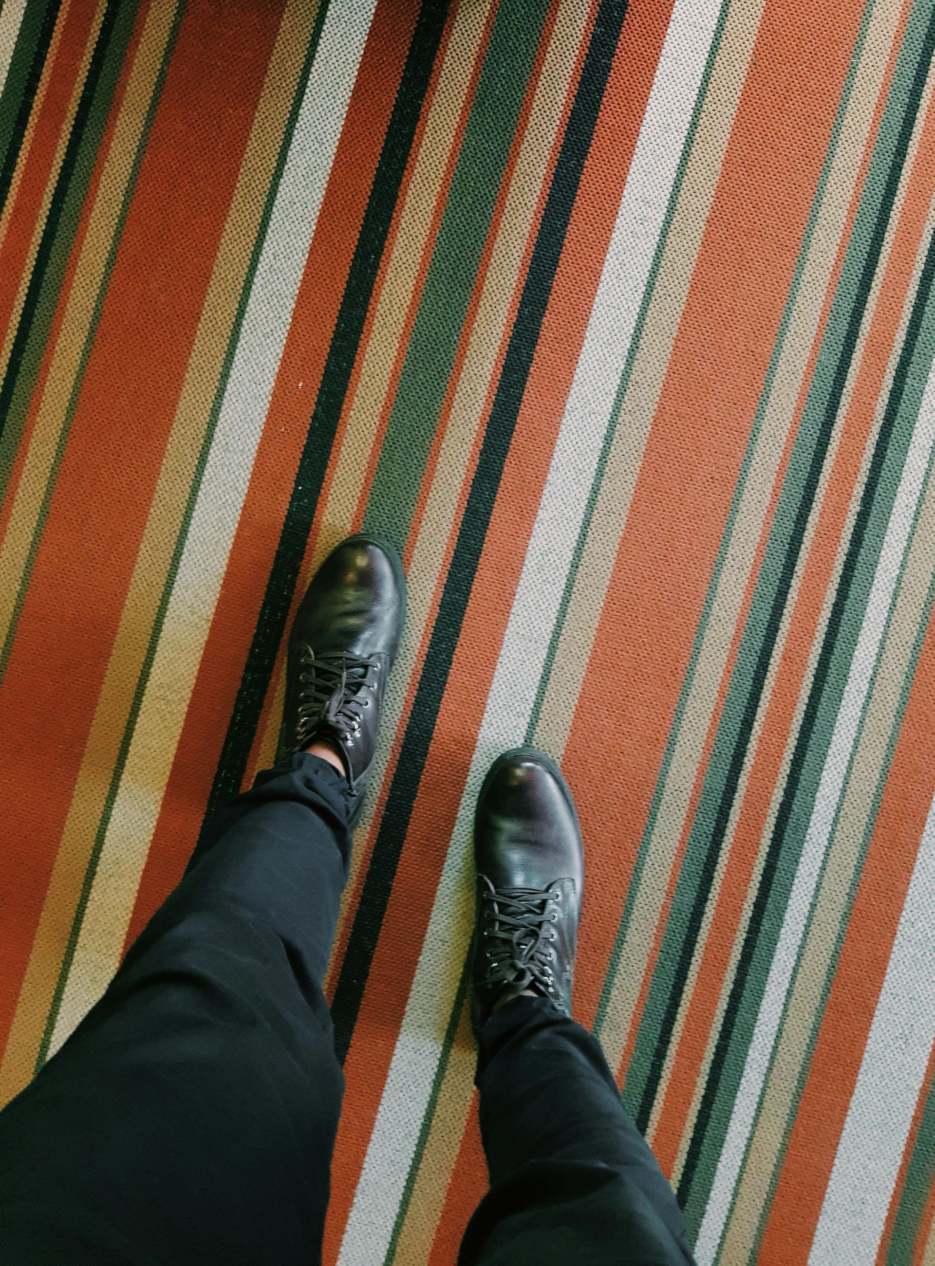 Person wearing black pants and boots standing on a striped carpet with orange, green, and white lines.