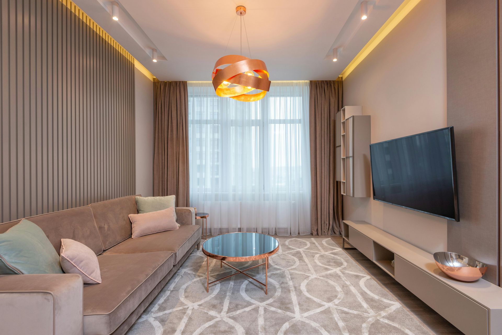 Living room with beige sofa, rug, TV, window with sheer curtains, and copper pendant light.