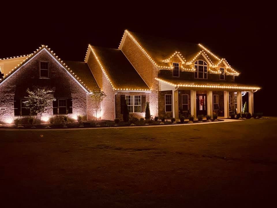 A large brick home illuminated with warm white Christmas lights outlining the roof and ground.