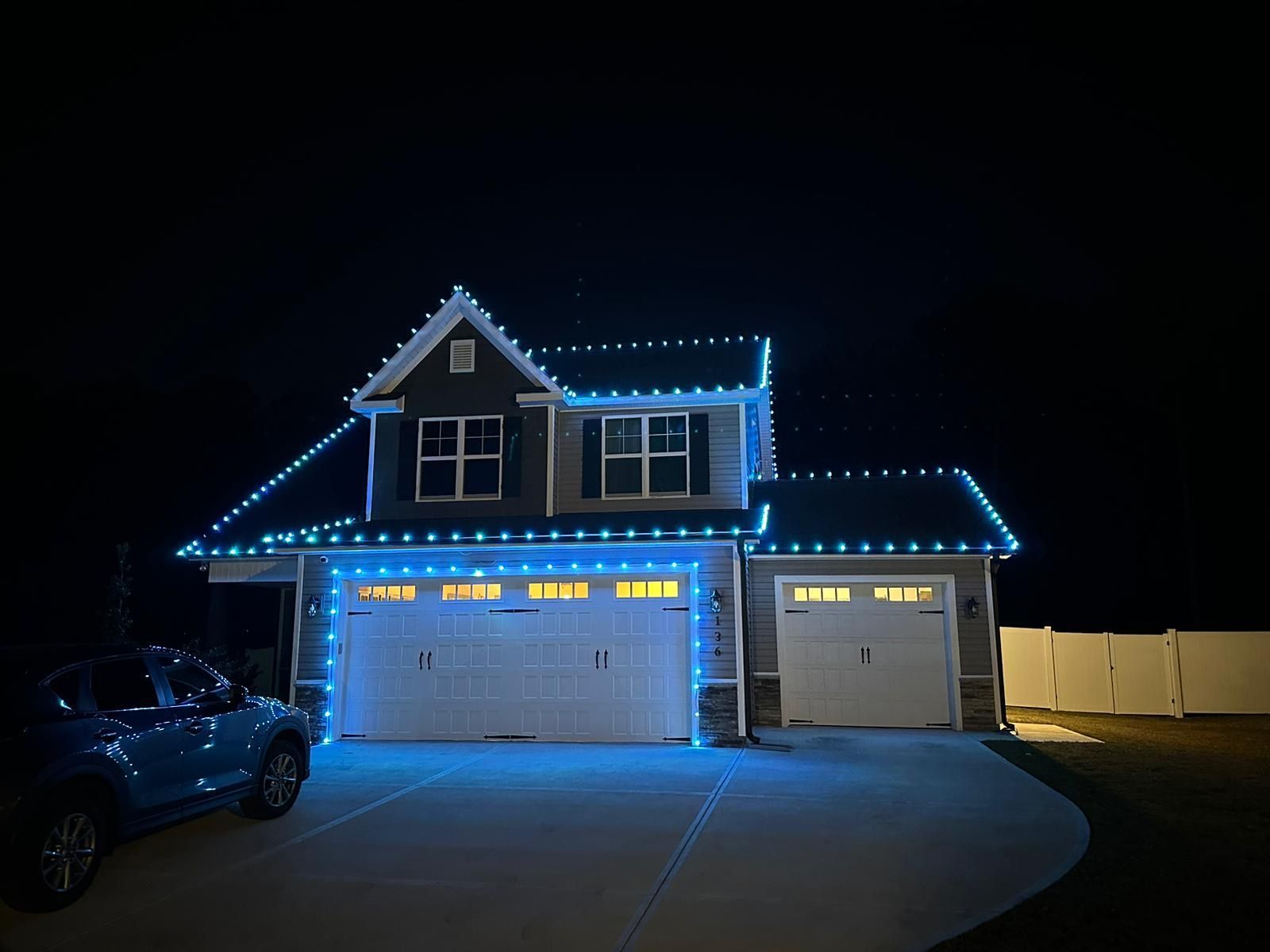 A house at night with blue Christmas lights outlining the roof and garage doors. A blue car is parked in the driveway.