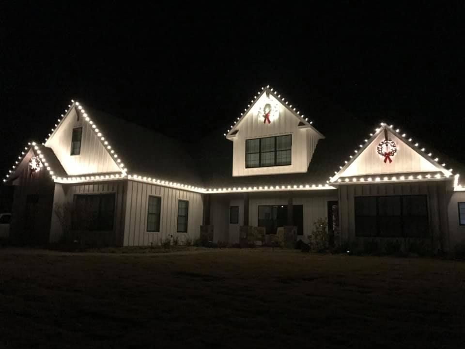 House with white siding, lit with white Christmas lights outlining the roof, wreaths on the peaks at night.