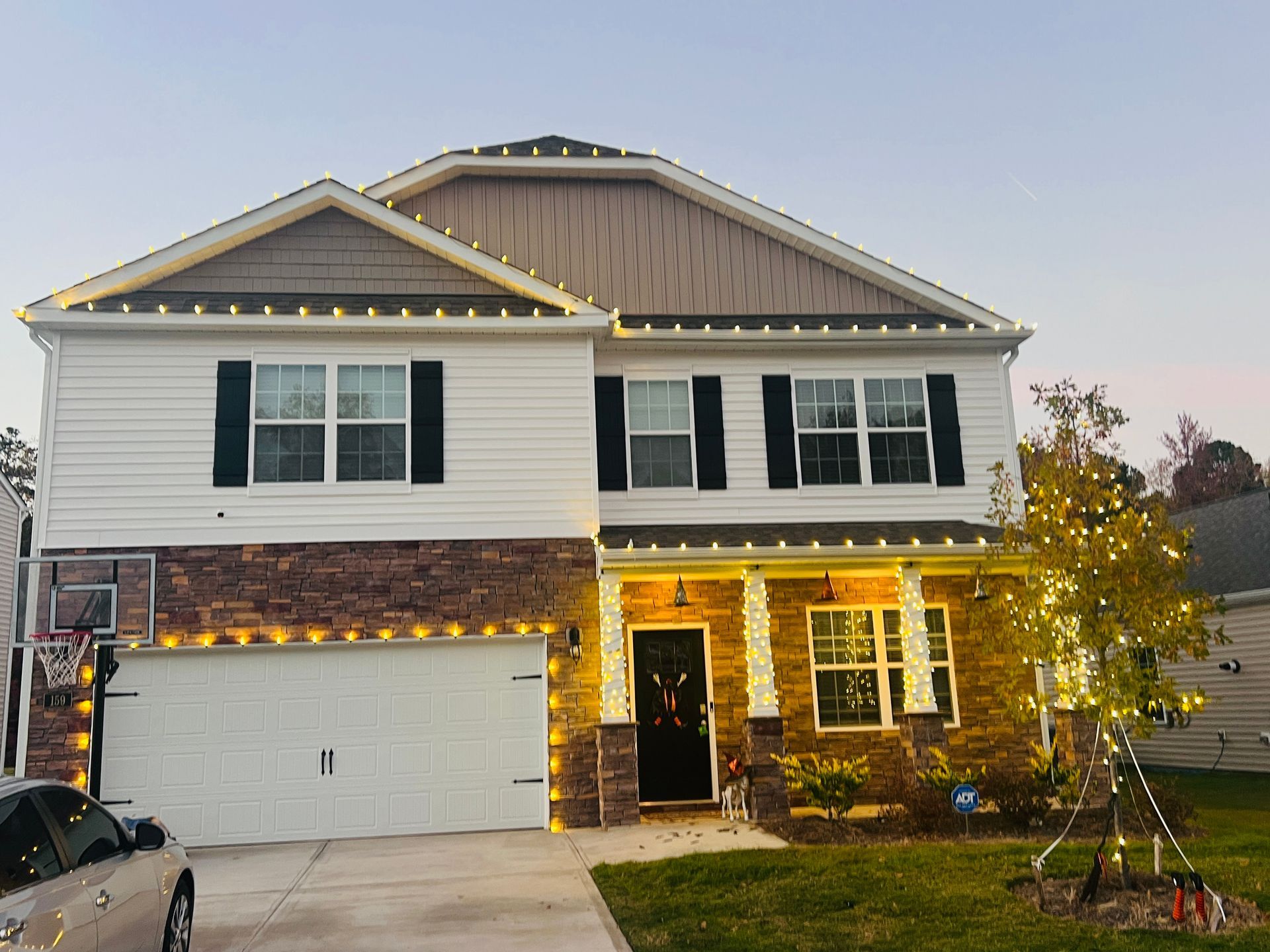 Two-story house decorated with string lights for the holidays. White siding, brown stone, black shutters.