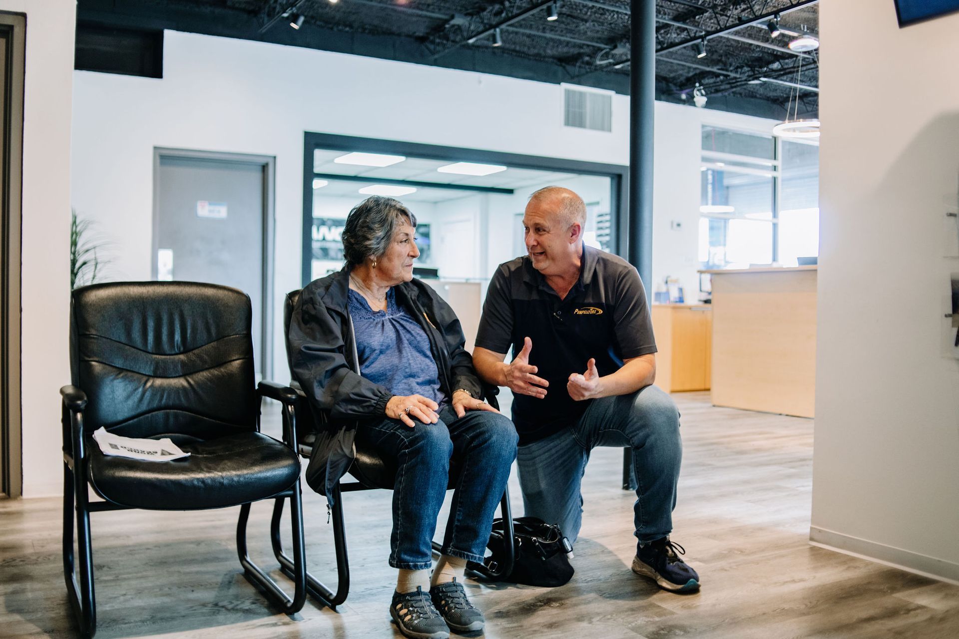 Two people talking on chairs in a bright office lounge, with another empty chair nearby.