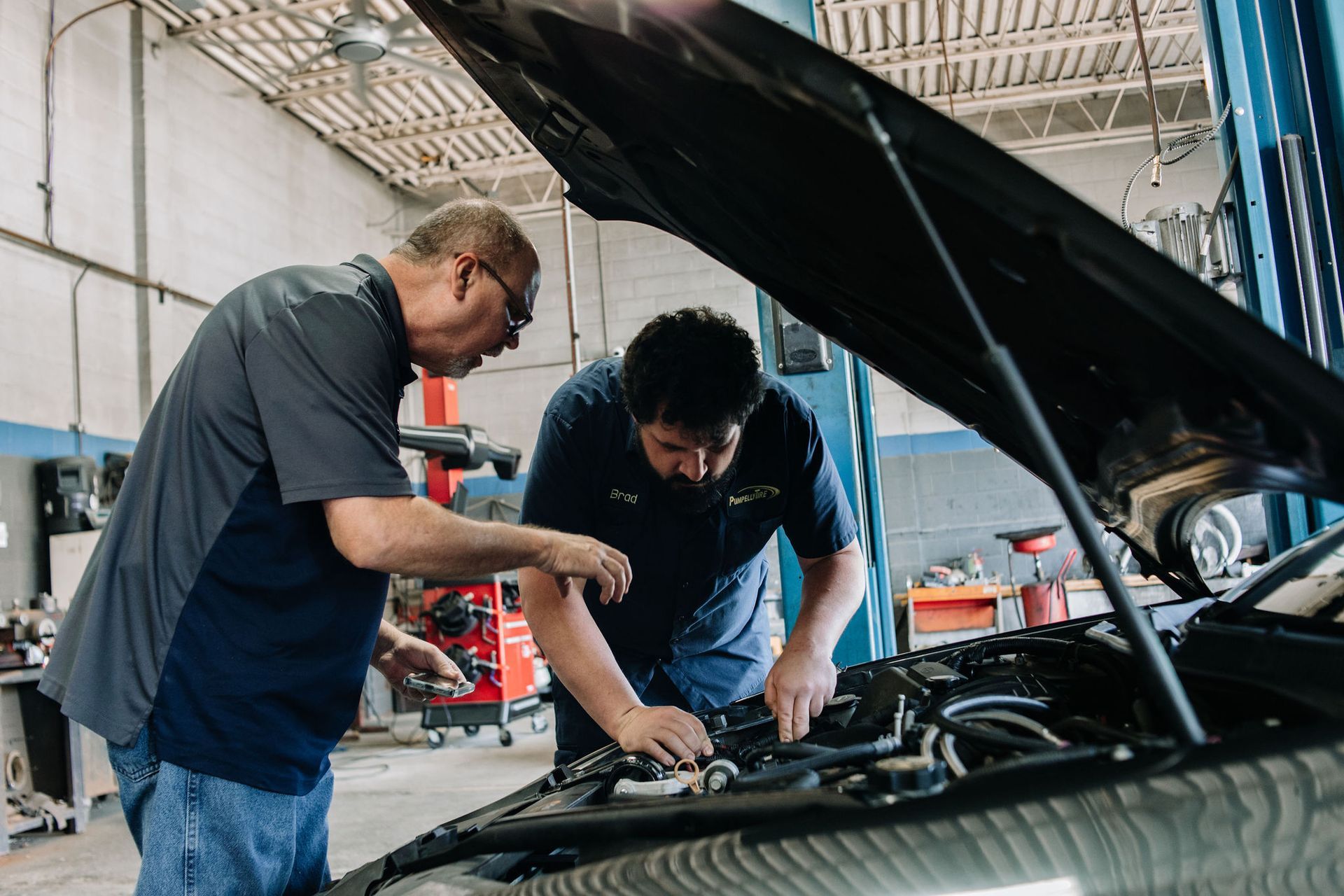 Two mechanics inspecting an open car engine in a garage, one pointing while the other works under the hood
