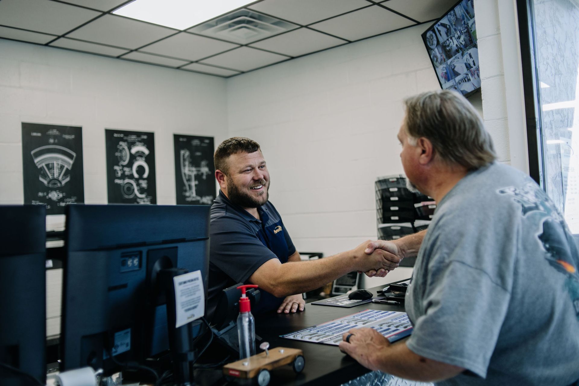 Two men shaking hands across a desk in an office, with framed posters on the wall.