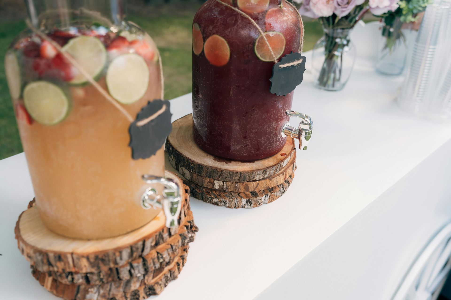 Two beverage dispensers on wood slabs with fruit-infused drinks, outdoors on a white table.
