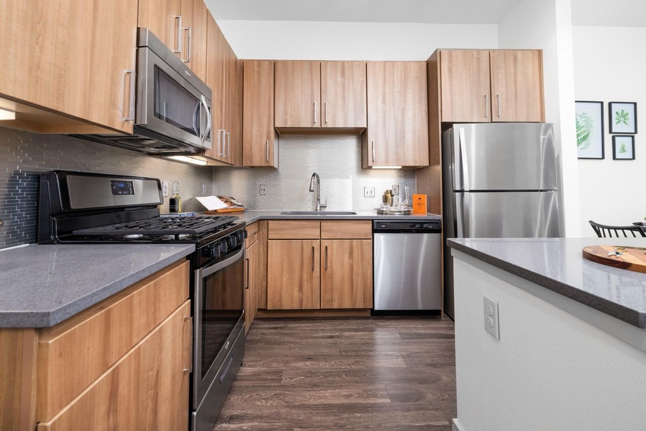 Modern apartment kitchen with wood cabinets, stainless steel appliances, and gray countertops.