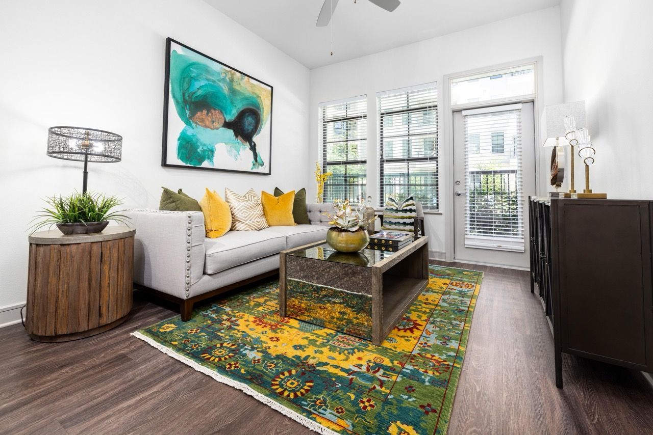 Bright living room in an apartment featuring a gray sofa, colorful rug, glass coffee table, and large windows.