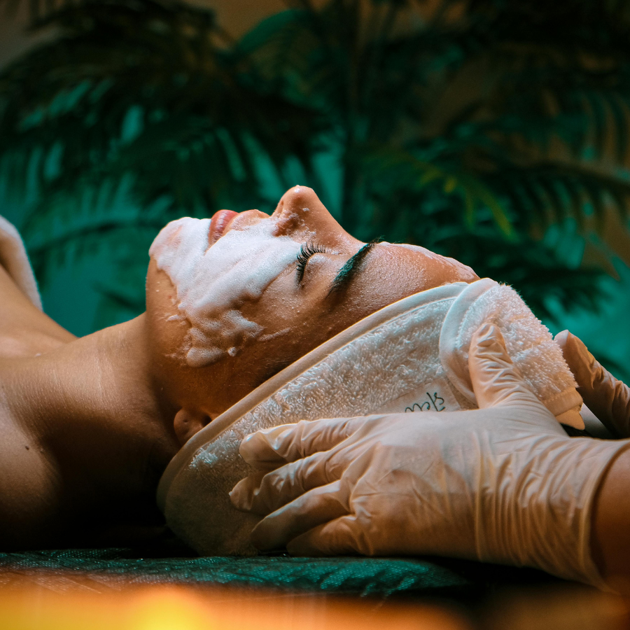 Person receiving a facial treatment at a spa; foam on face, gloved hands, palm fronds in background.