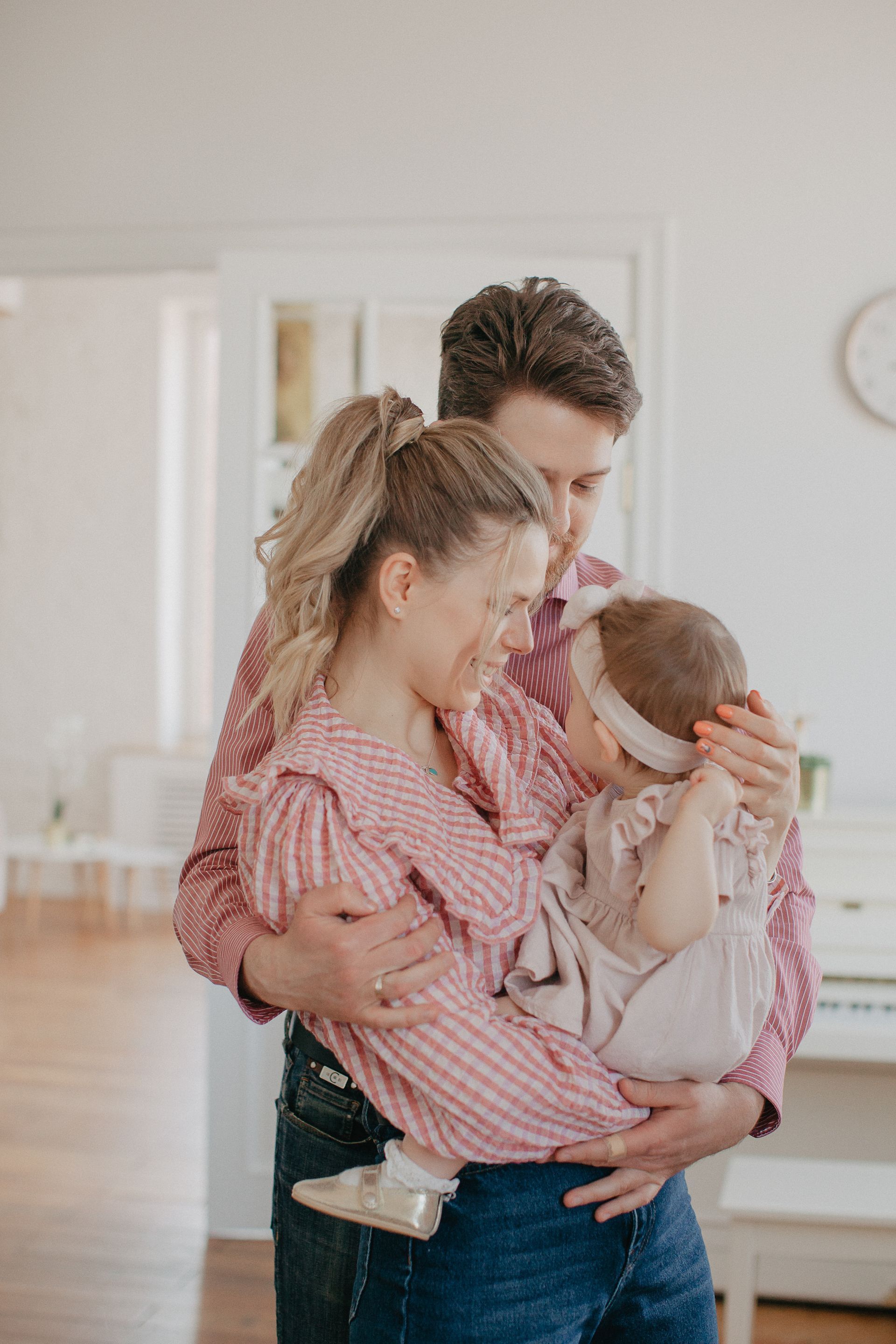 Family indoors; parents holding a baby in a pink-striped shirt, smiling.