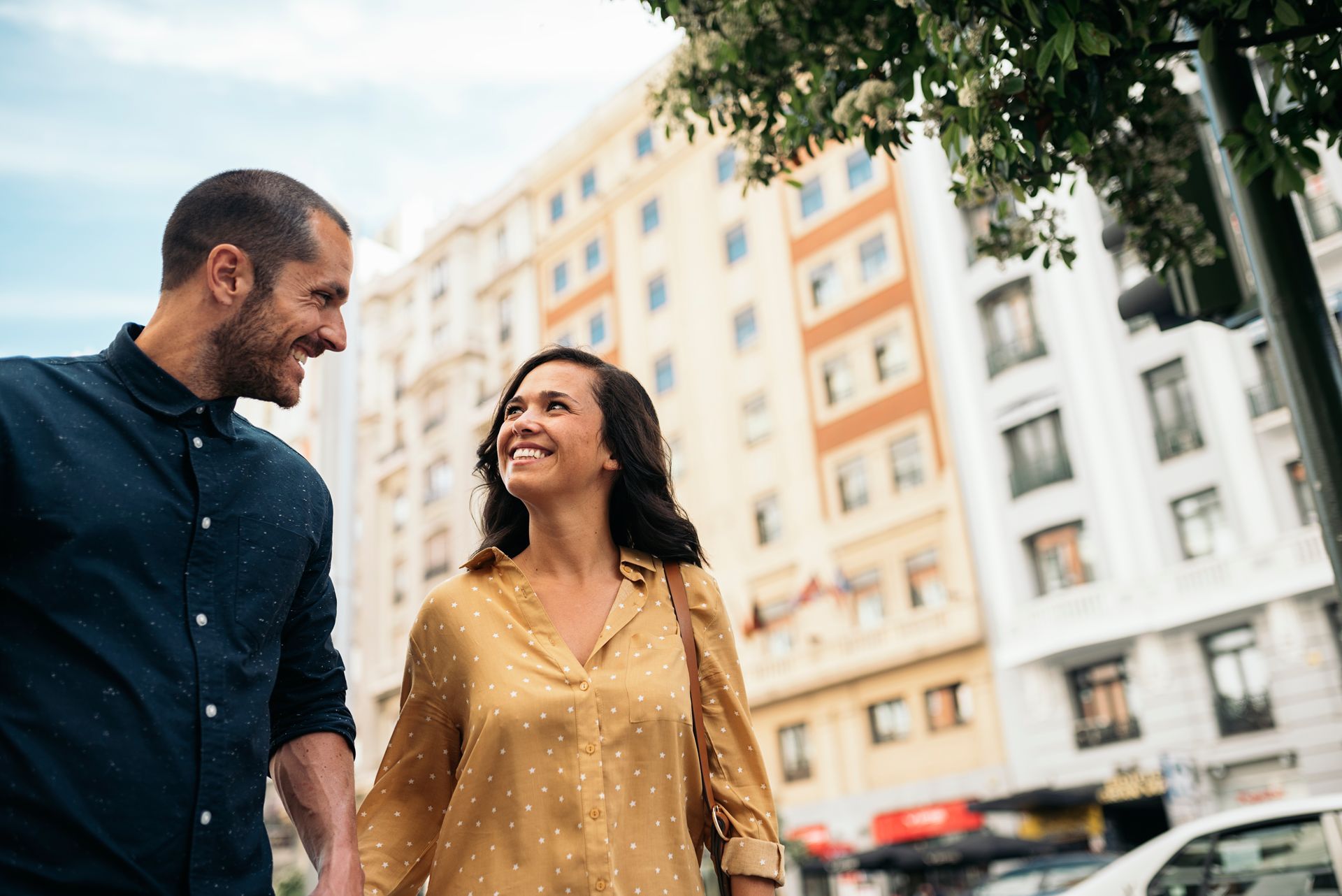Couple walking and holding hands, smiling at each other in front of a city building.