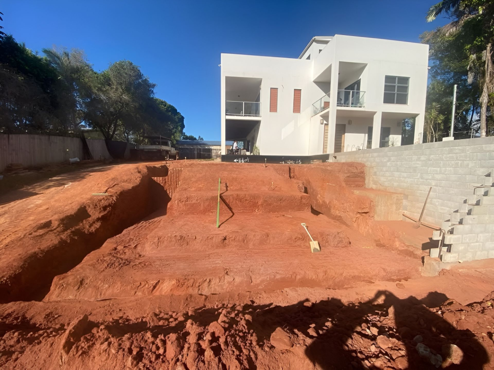 Construction site with a house in the background, with red dirt and retaining wall in progress.— BC Earthmoving & Civil Construction in East Barron QLD