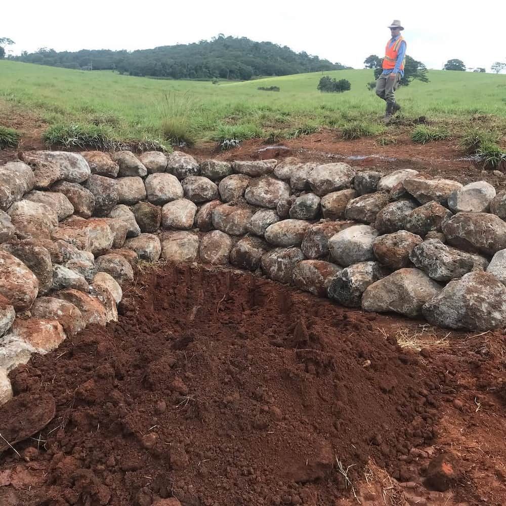 A Stone Retaining Wall Being Constructed in a Grassy Field — BC Earthmoving & Civil Construction in East Barron QLD
