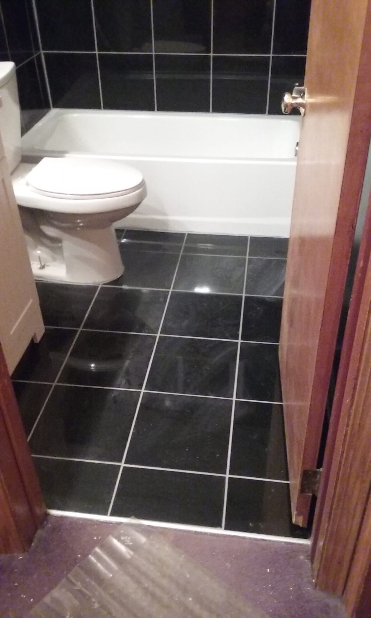 A view looking into a small bathroom featuring black square floor and wall tiles, a white toilet, and a white bathtub.