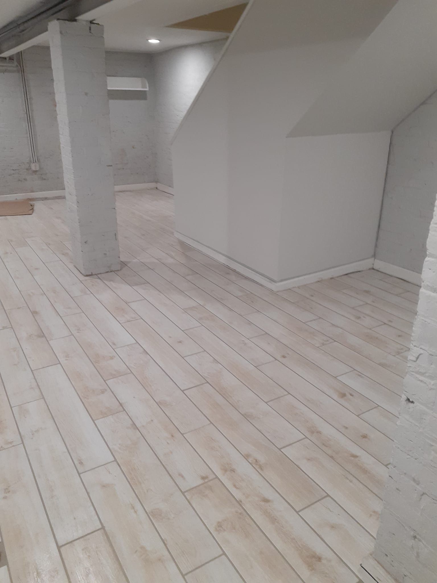 A room with light wood-look tile floors, a white structural column, and a staircase wall in a basement-style interior.