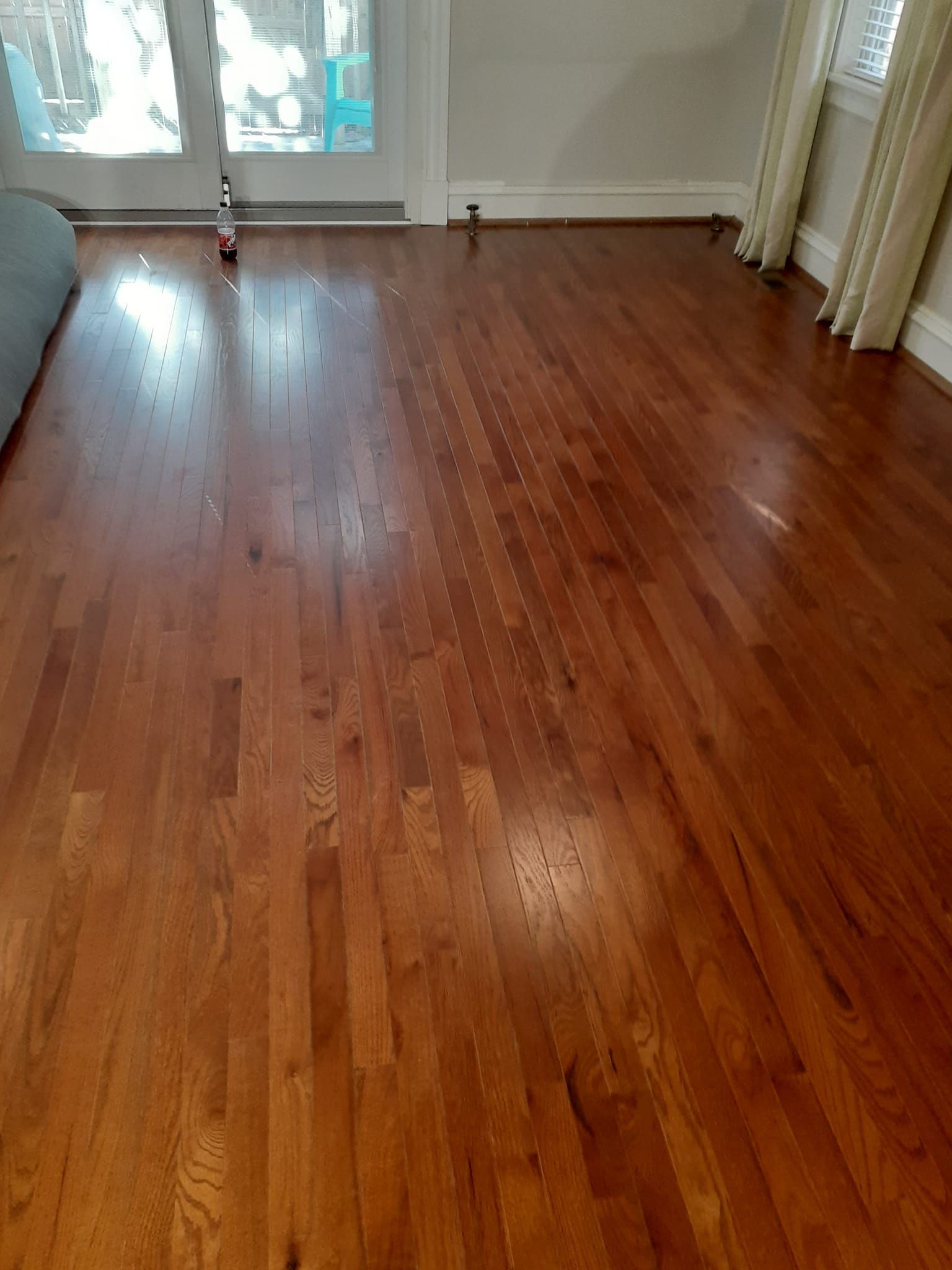 A room with polished, medium-toned hardwood flooring leading toward glass doors and white curtains.
