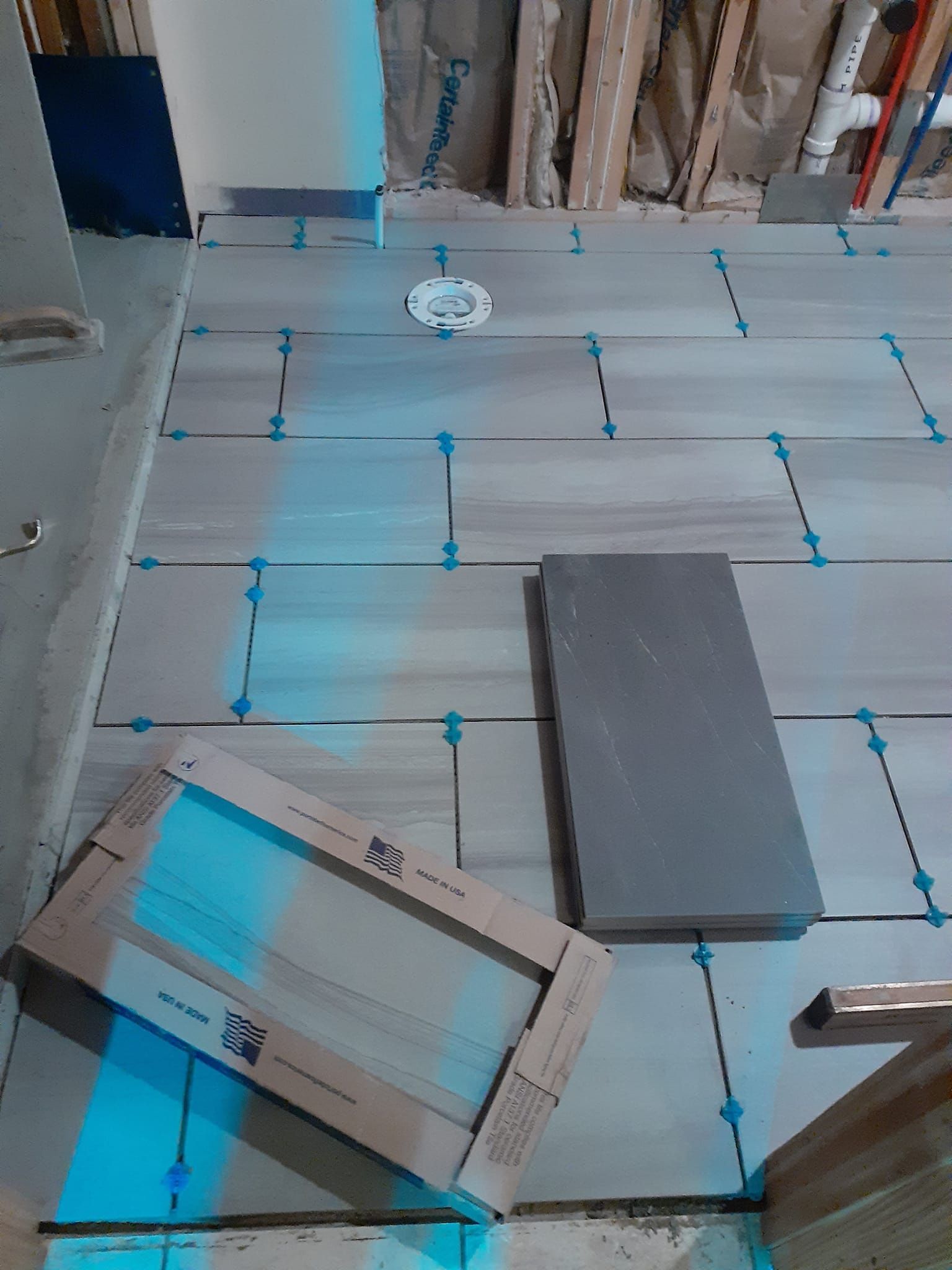 Tiles laid on a floor with spacers, a laser level line, a box of tiles, and a plumbing flange in a room under renovation.