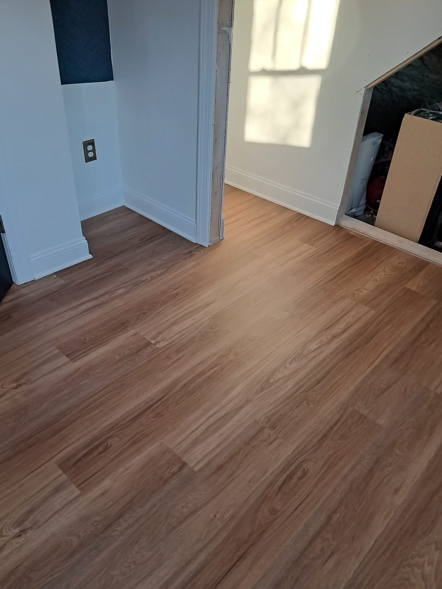 A room with light brown wood-look flooring, white walls, and a doorway leading to a sunlit space with an angled wall nook.