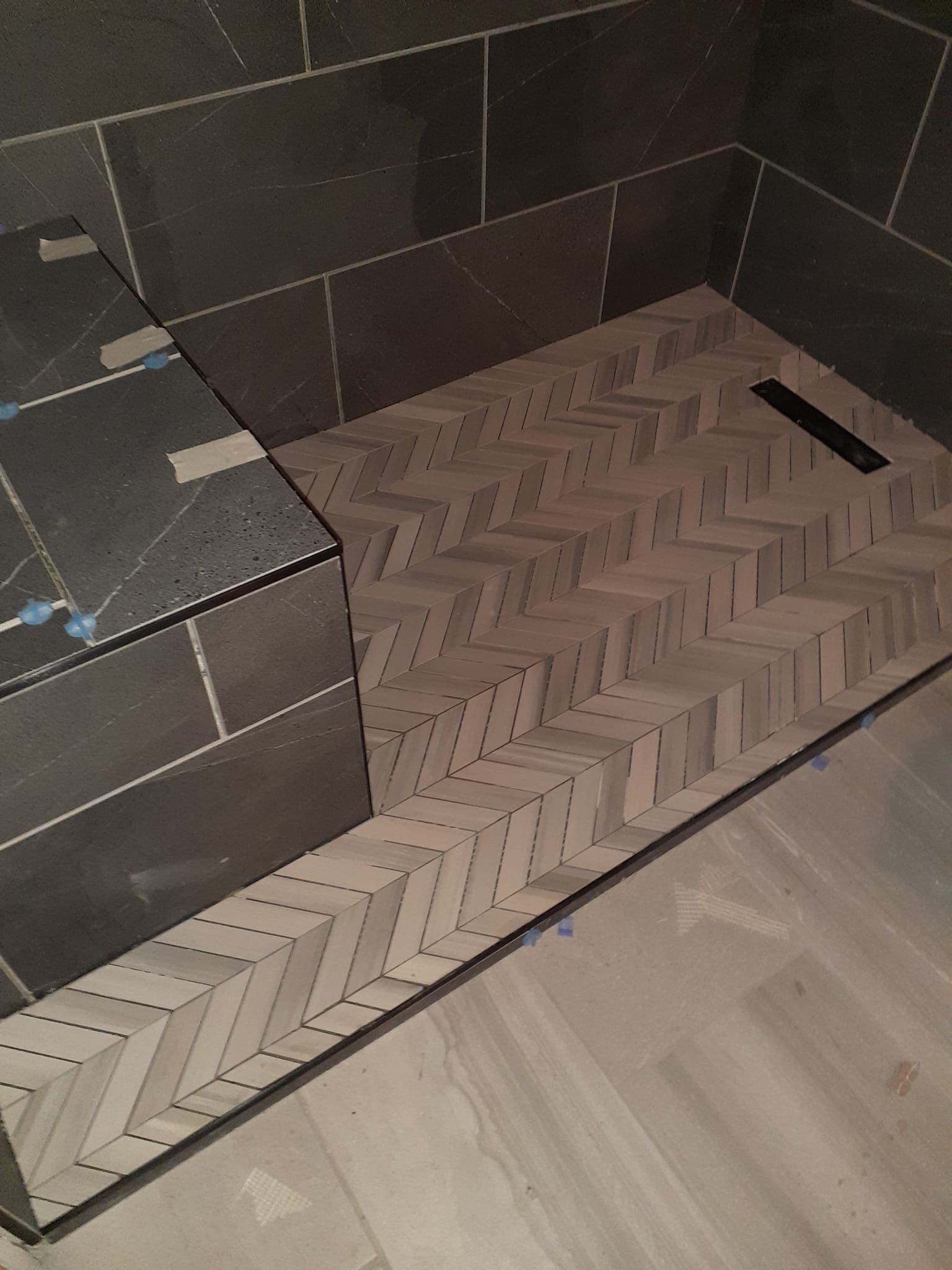 A shower floor featuring light-colored, herringbone-patterned tile, a dark bench, a linear drain, and large gray wall tiles.