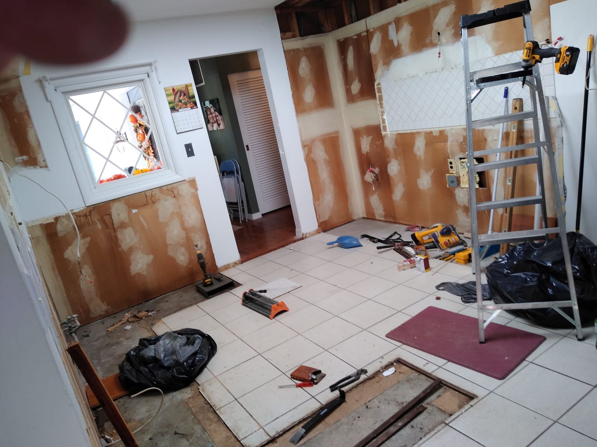 An interior room under renovation with exposed wall panels, tiled floor, a ladder, and construction tools scattered about.