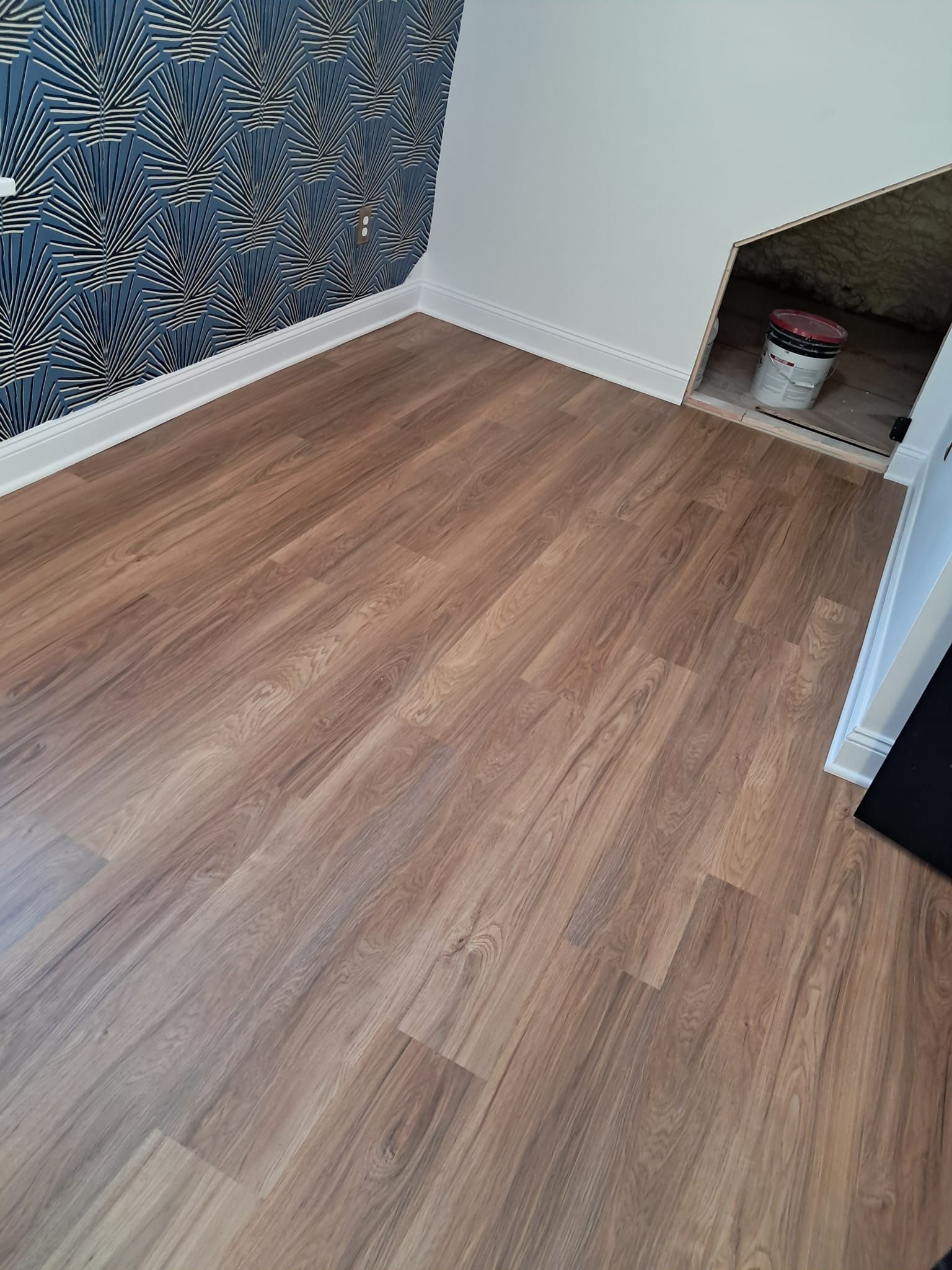A room featuring light brown wood-look flooring, a patterned blue accent wall, white trim, and a small, open crawl space.