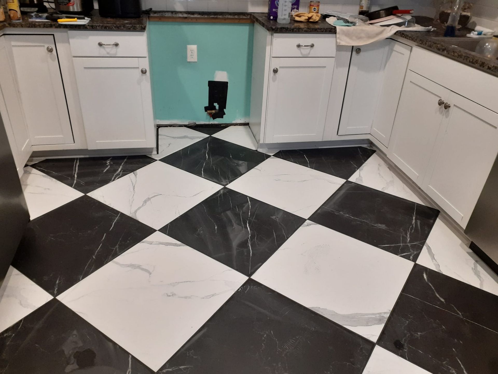 A kitchen floor with a black and white diamond-patterned tile design, flanked by white cabinets and a teal wall section.
