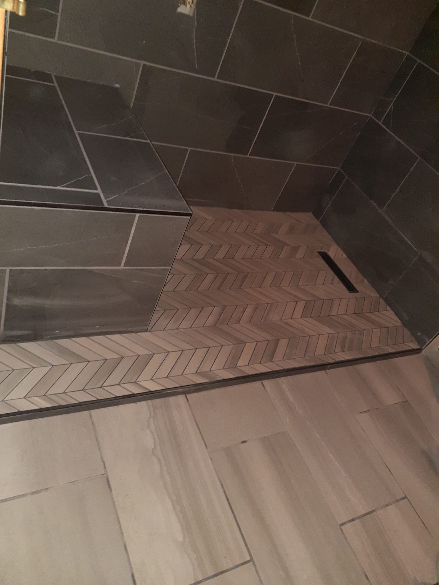A walk-in shower with dark grey wall tiles, a built-in tiled bench, and a light-colored herringbone floor with a drain.