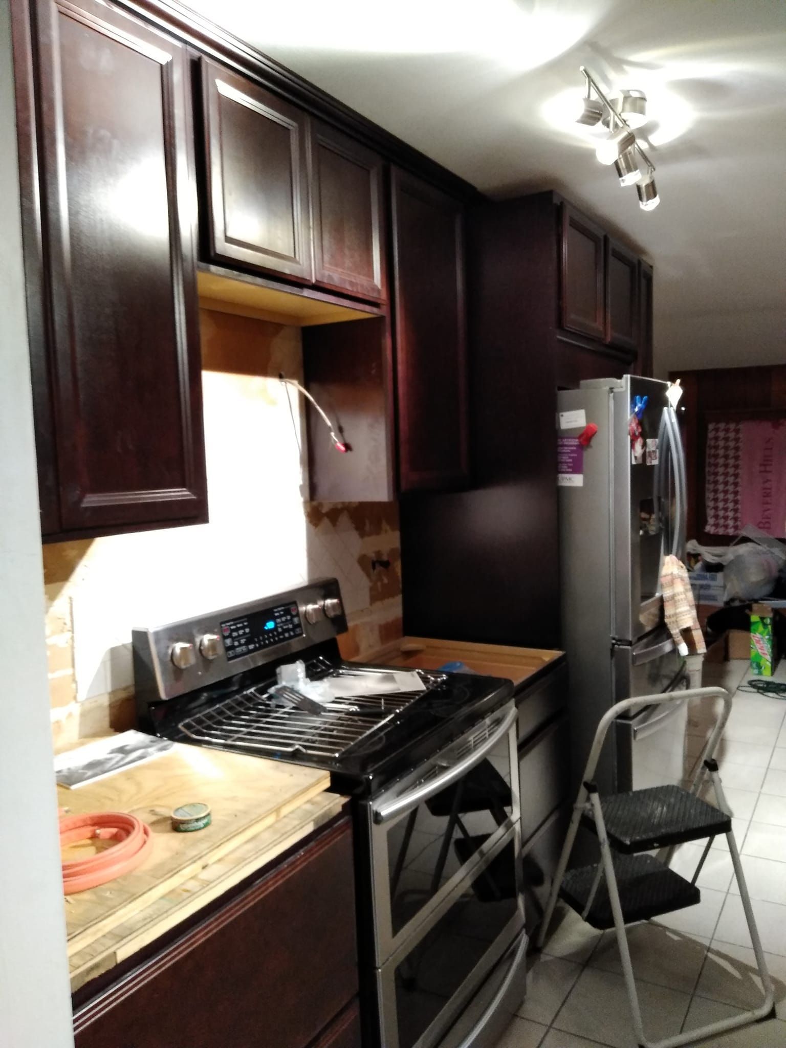 A kitchen under renovation with dark wood cabinets, a stainless steel stove, and a folding step ladder on a tiled floor.