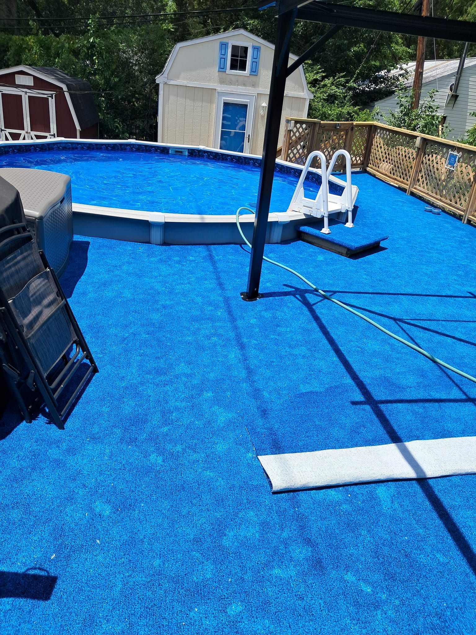 An above-ground swimming pool on a deck with vibrant blue carpeting, a white ladder, and two small sheds in the background.
